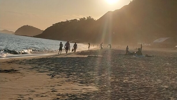 Une plage où l’on voit des gens se promener et s’asseoir, le soleil se couche à l’arrière-plan et des collines se dessinent à l’horizon.