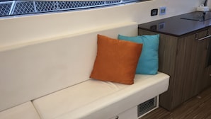 A boat interior with a cushioned bench, a small kitchen area, and a window with a mesh screen.