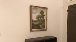 Un meuble en bois sombre, doté de tiroirs, est adossé à un mur uni. Au-dessus, on aperçoit un tableau représentant un paysage, encadré. Une porte fermée se trouve à droite.