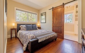 A bedroom with a large bed, two bedside tables with lamps, a wooden door, and a view of trees through the window.