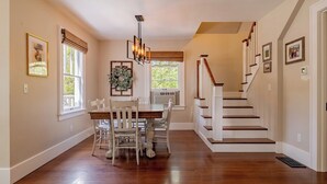 Un comedor con mesa y sillas de madera, una araña y una escalera con barandillas blancas.