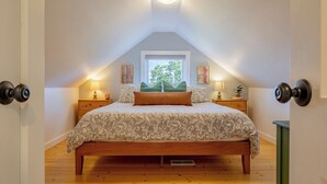 Un dormitorio con techo inclinado, una cama con ropa de cama estampada, dos lámparas de noche, una ventana con vistas a follaje y una puerta con pomo redondo.