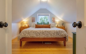 Un dormitorio con techo inclinado, una cama con ropa de cama estampada, dos lámparas de noche, una ventana con vistas a follaje y una puerta con pomo redondo.