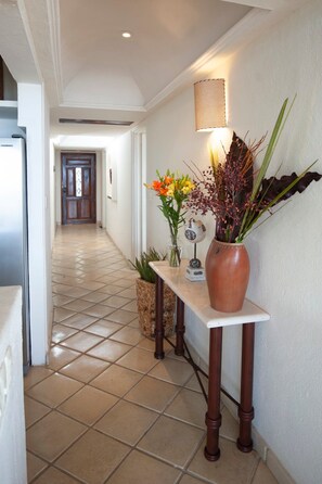 A hallway with a tiled floor, a wooden table with a vase of flowers, and a wall-mounted lamp. - Ocean Front/Heated Pool, 6 Adults+ 2 kids under 12 (Puerto Vallarta)