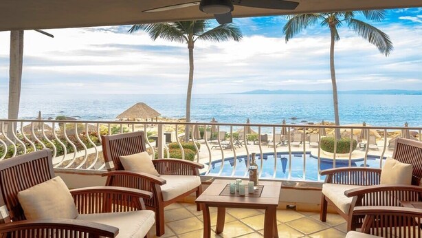 Un balcon avec des chaises et une table en bois, donnant sur une piscine et la mer.