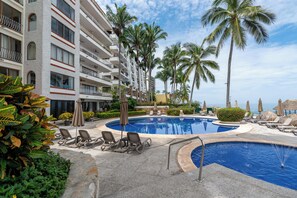 A resort with a swimming pool, palm trees, and a view of the ocean. - Ocean Front/Heated Pool, 6 Adults+ 2 kids under 12 (Puerto Vallarta)
