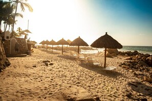 A beach with thatched umbrellas, lounge chairs, and a rocky shoreline. - Ocean Front/Heated Pool, 6 Adults+ 2 kids under 12 (Puerto Vallarta)