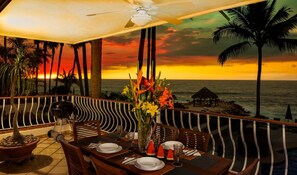 Een eethoek met uitzicht op de oceaan bij zonsondergang, met een gedekte tafel voor twee, een bloevas en een grill.