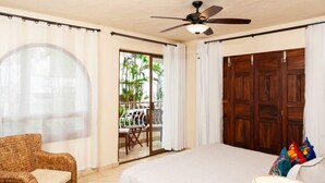 Une chambre avec un lit, une chaise en osier, un ventilateur de plafond et une vue sur un espace extérieur aménagé avec des chaises et des plantes.