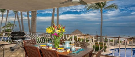 A balcony with a table set for dining, overlooking a pool and the ocean.
