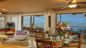 Un vaste espace de vie comprenant une salle à manger avec une table dressée pour six personnes, un coin salon confortable et un balcon offrant une vue sur la mer.