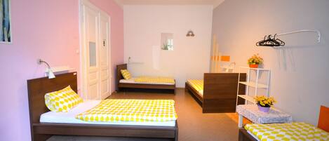 A room with three beds, a small seating area, a shelf with a plant, and a wall-mounted hanger.