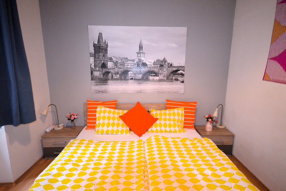 A bedroom with a bed featuring a yellow and white patterned bedspread, a large orange pillow, and a framed black and white cityscape picture on the wall.