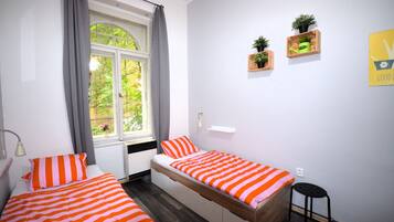 Una habitación con dos camas, una ventana con cortinas, un taburete pequeño y dos macetas colgantes.