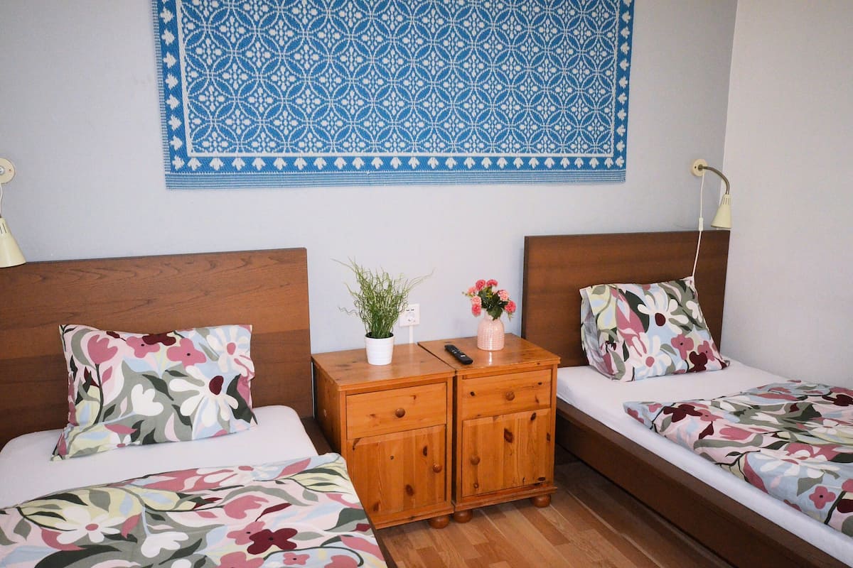 A double bed with floral bedding, a wooden nightstand with a plant and a vase, and a blue patterned tapestry on the wall.