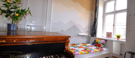 A room with a piano, a bed with a colorful quilt, a window with a view, and a plant on the windowsill.
