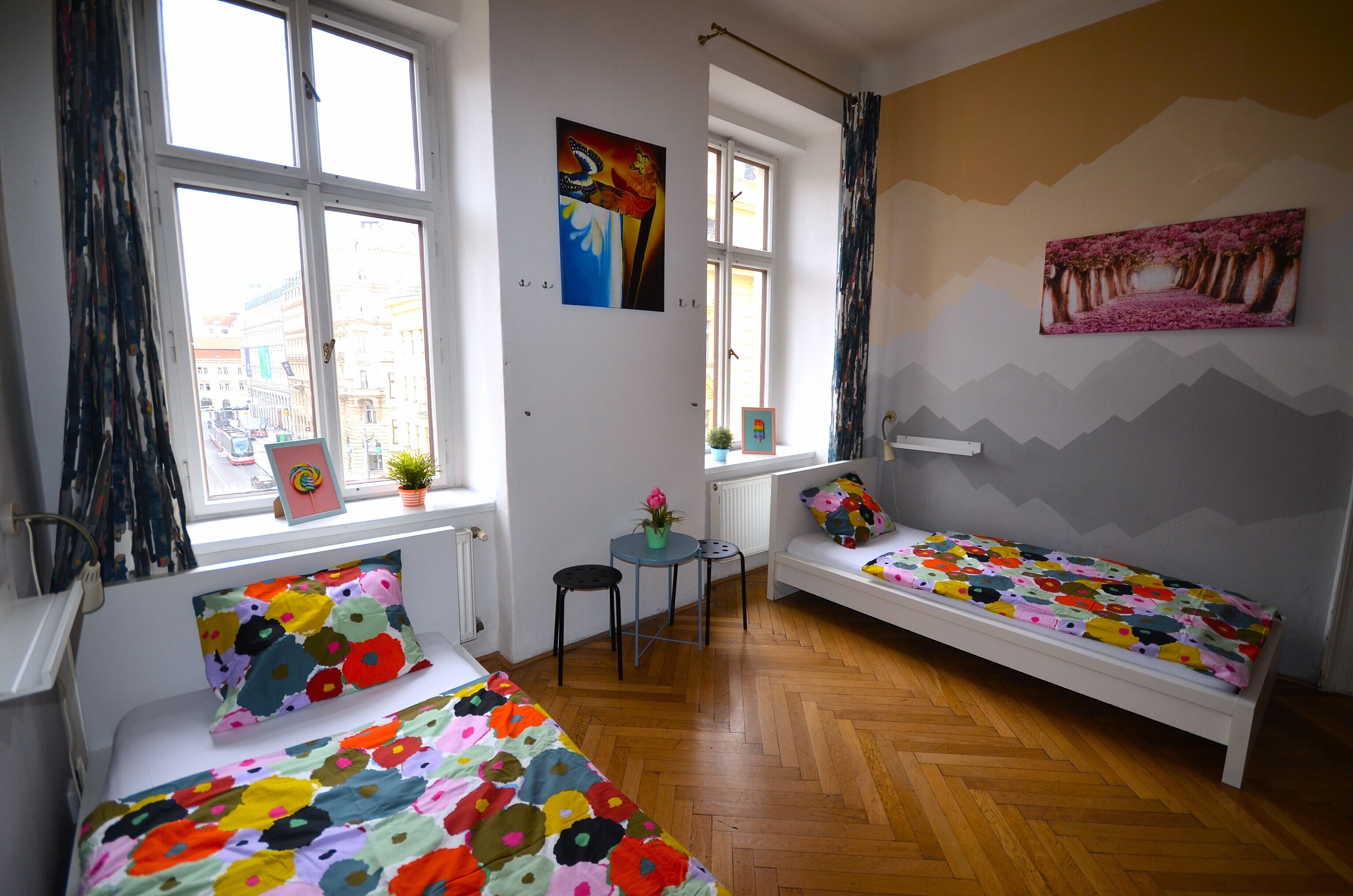 A room with two single beds, a small table, a potted plant, and a window with a view of buildings.