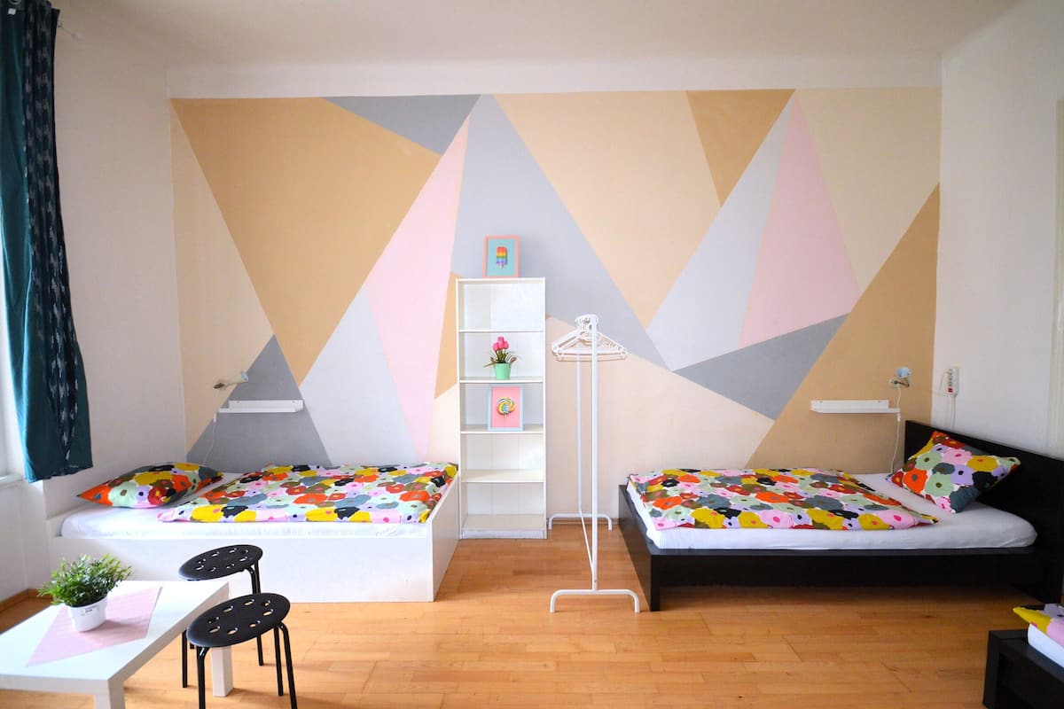 A room with two beds, a small table with a chair, a white shelf unit, and a floor lamp.