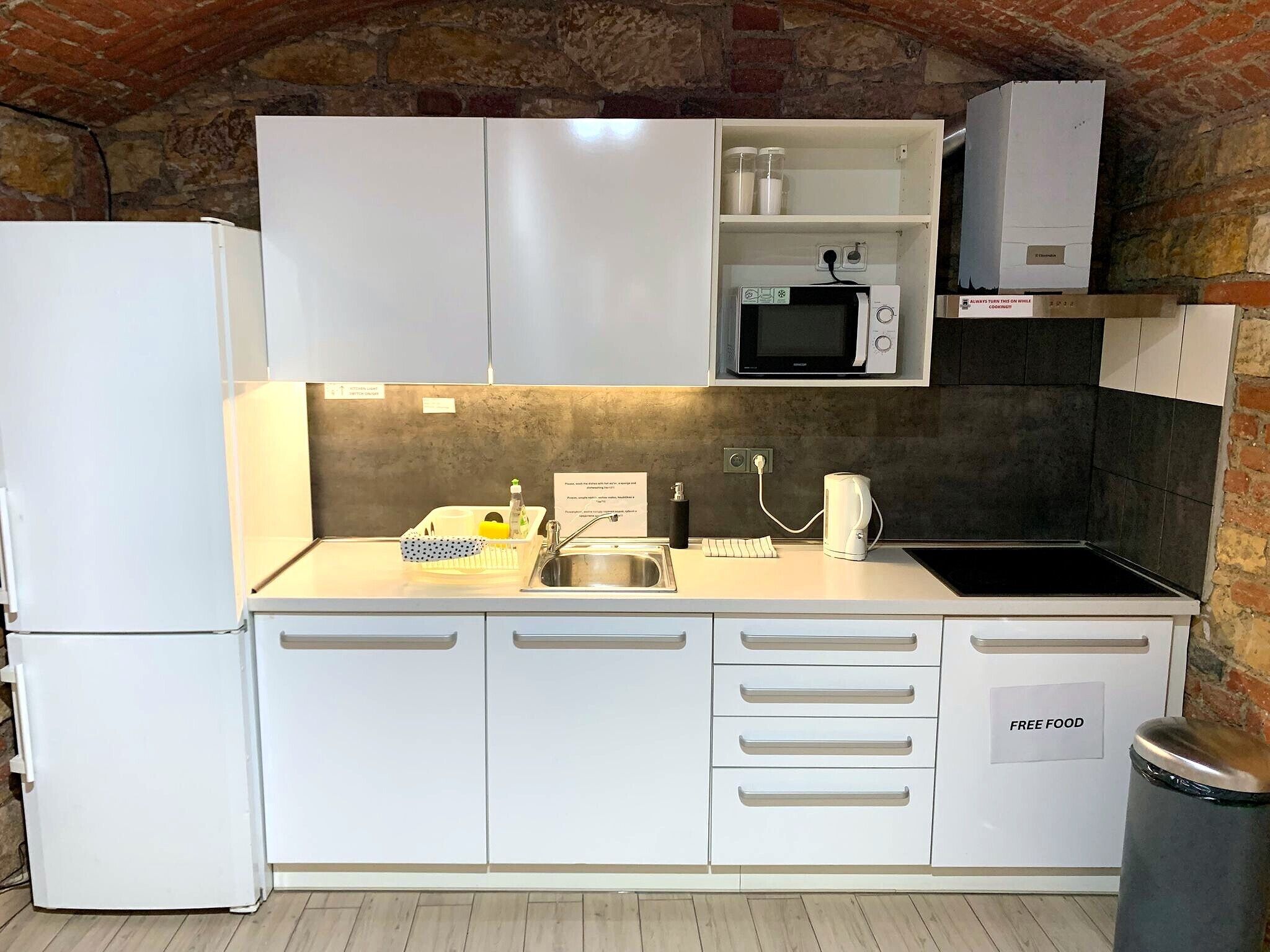 Una cocina pequeña con gabinetes blancos, un microondas, un lavamanos y un cubo de basura.