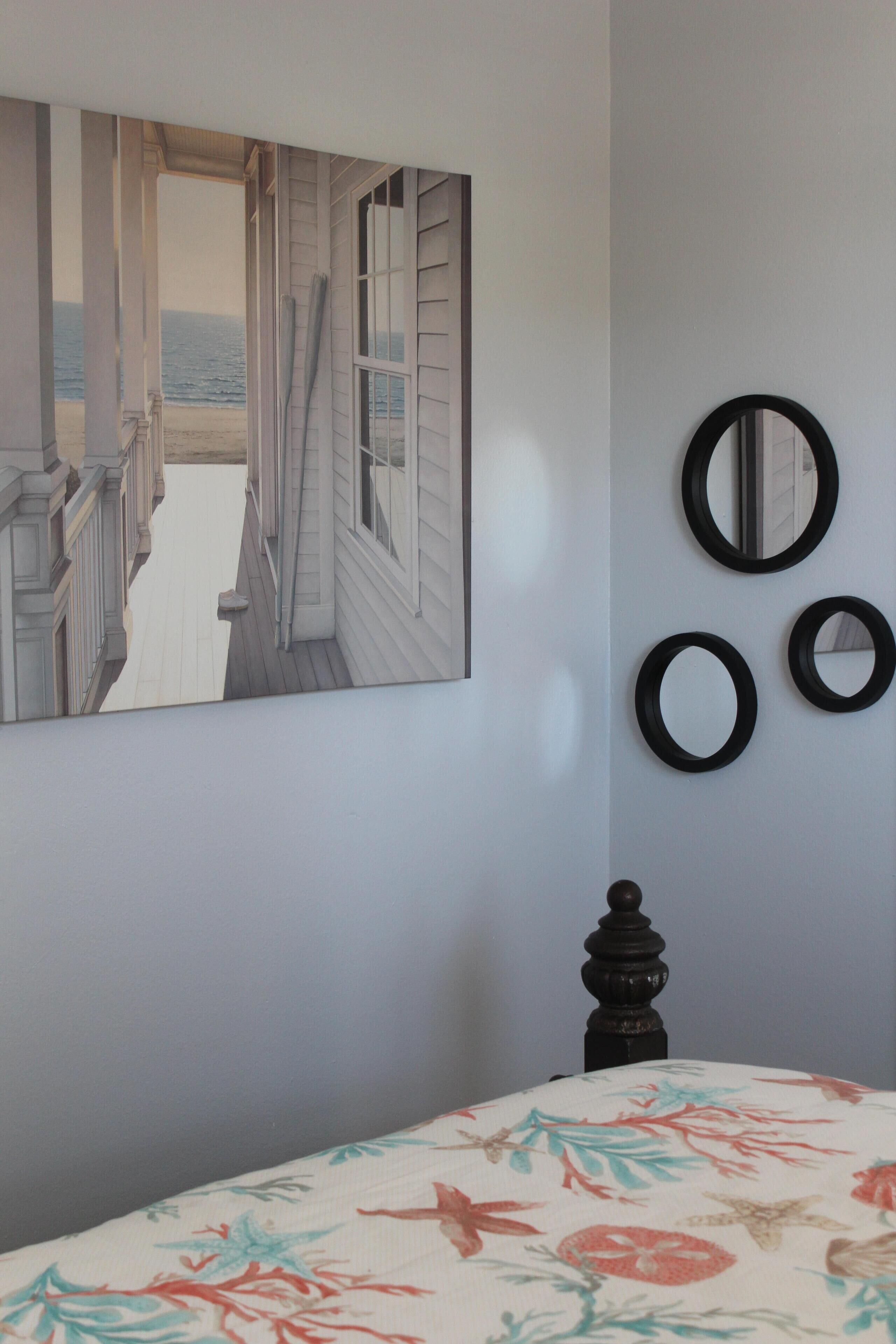 A bedroom with a bed, a painting of a coastal scene, and three wall-mounted mirrors.