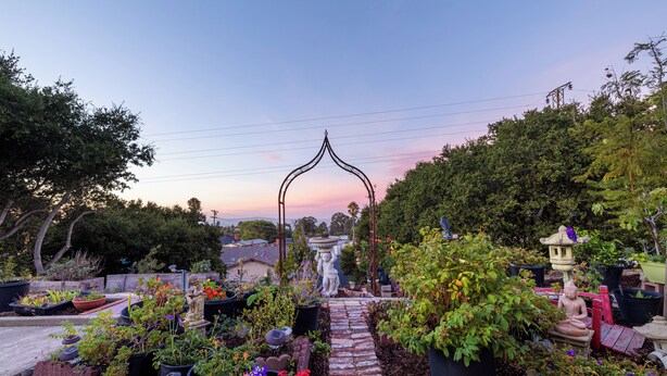 Un sendero de jardín conduce a un pabellón con una estatua, rodeado de macetas con plantas y una estatua de una figura en meditación.