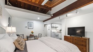 Un dormitorio con cama, cómoda, televisor y una kitchenette al fondo.