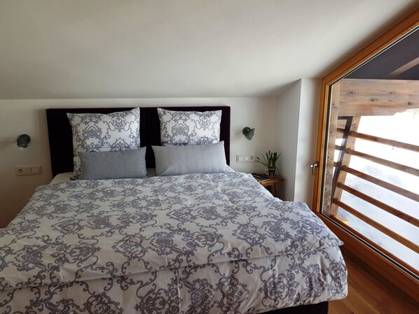 A bedroom with a large bed, two wall-mounted nightstands, a potted plant, and a wooden-framed window. - Kaiserwetter vacation apartment, 80sqm, with balcony and high-quality furnishings (Reit im Winkl)