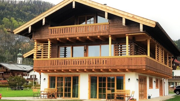 Une maison en bois à deux étages, dotée d’un balcon, de grandes fenêtres et d’une terrasse aménagée avec du mobilier d’extérieur.