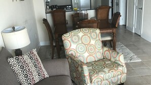 Un salon avec un canapé gris, un fauteuil à motifs et une petite table ronde sur laquelle repose un magazine. Une lampe et une décoration en forme d’étoile ornent le mur.