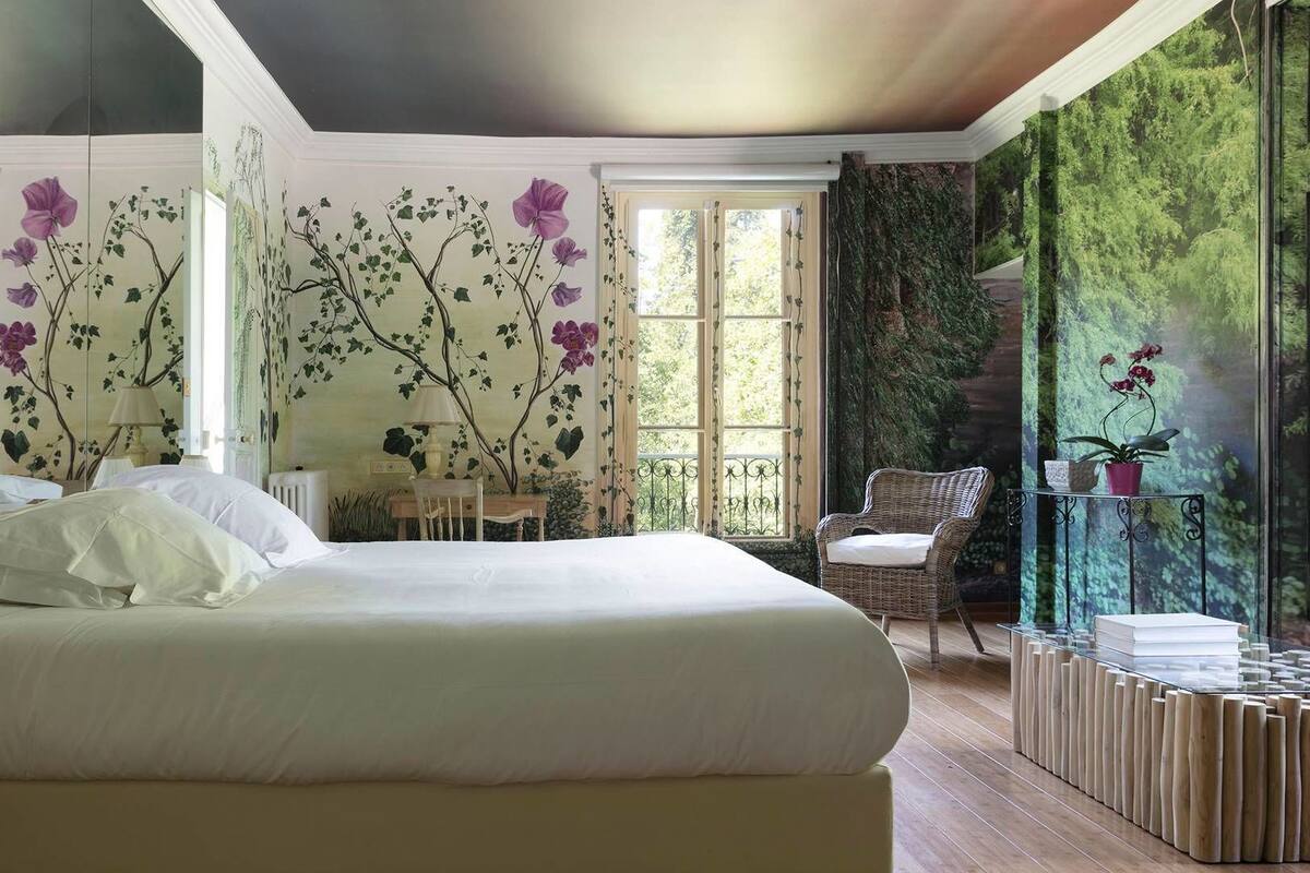 Un dormitorio con una cama grande, una silla, una mesita y una ventana con cortinas.