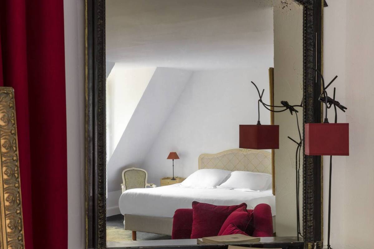 Habitación de hotel con una cama, una silla, una mesita de noche y un espejo que refleja la habitación.