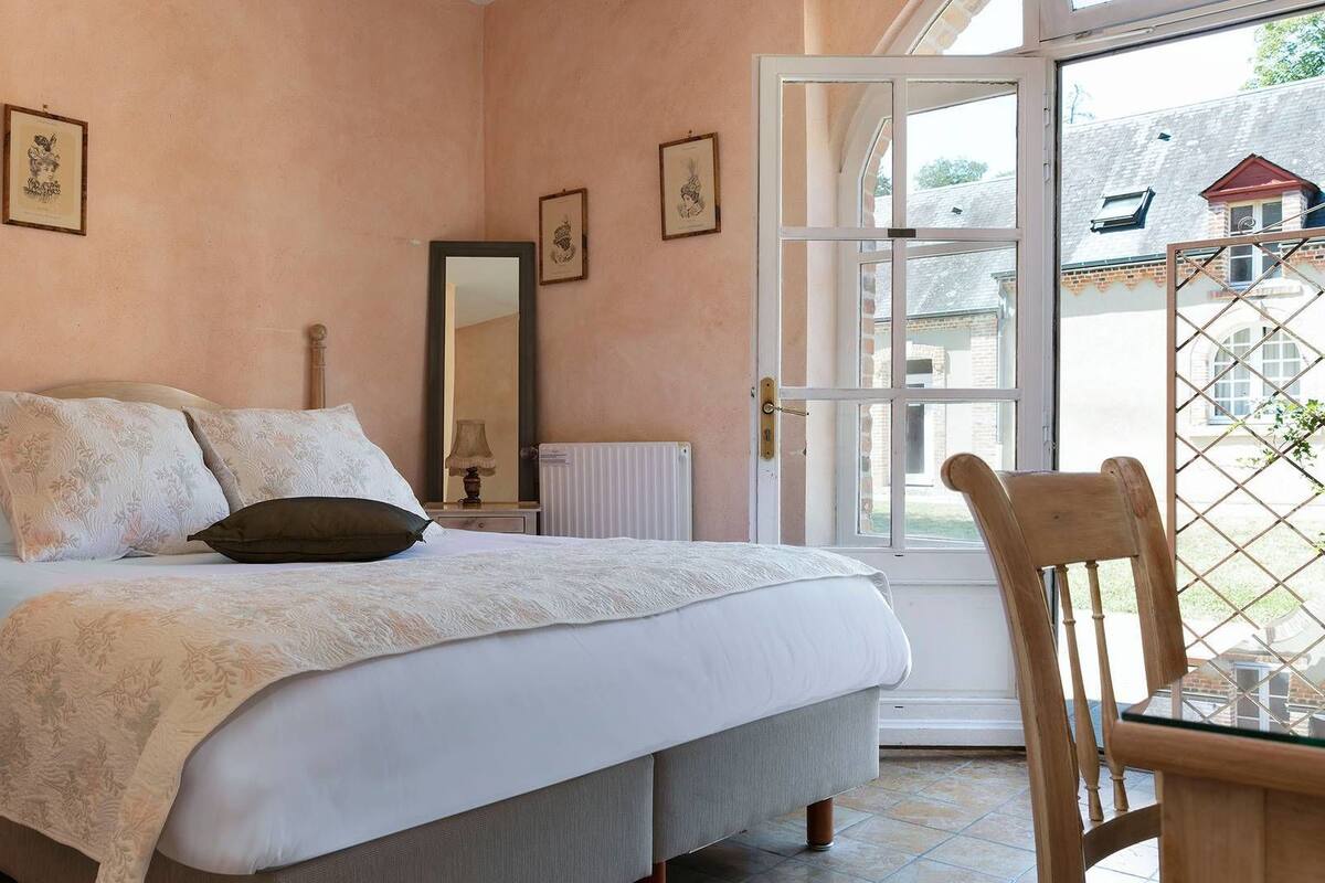 Un dormitorio con cama, una silla, una mesa, un espejo y una ventana.