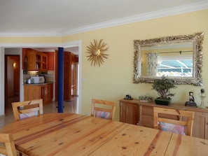 A dining area with a wooden table and chairs, a kitchen in the background, a decorative sunburst wall piece, and a mirror with an ornate frame.