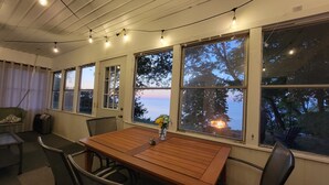 Une pièce avec une table et des chaises en bois, de grandes fenêtres décorées de motifs en forme d’étoiles et une vue sur la mer au crépuscule.