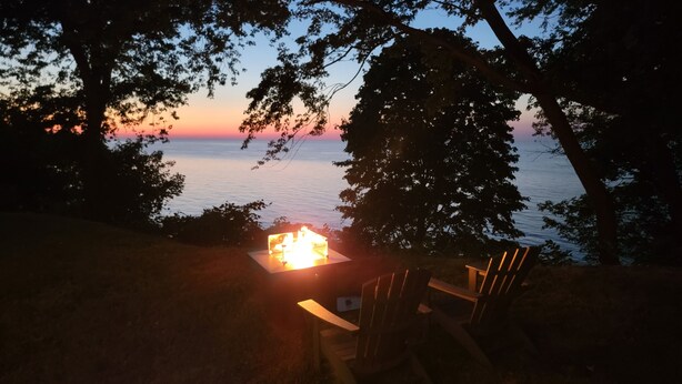 Un feu de camp avec une table et des chaises, installé dans un cadre naturel, entouré d’arbres et offrant une vue sur une étendue d’eau au coucher du soleil.