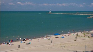 Une plage où des gens se prélassent au soleil et se baignent, un phare à l’horizon et un ciel dégagé.