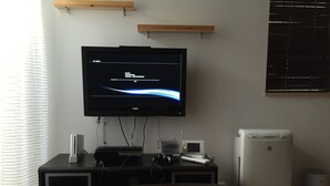 Una habitación con un televisor de pantalla plana montado en la pared, una mesita con dispositivos electrónicos y un electrodoméstico blanco en el suelo.
