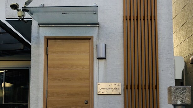 Una puerta de madera con un picaporte de metal, un letrero que dice "KatSuragawa House" y un escalón que lleva a la entrada.