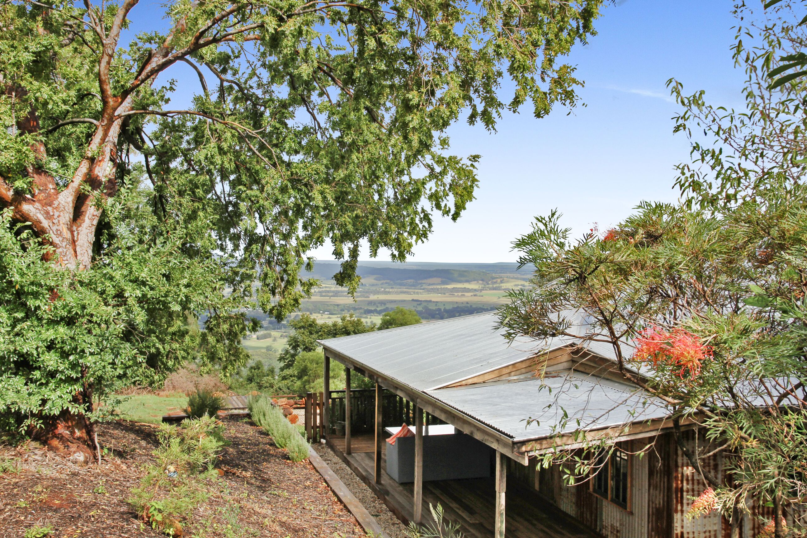 A rustic wooden building with a metal roof, surrounded by lush greenery and a view of distant hills.