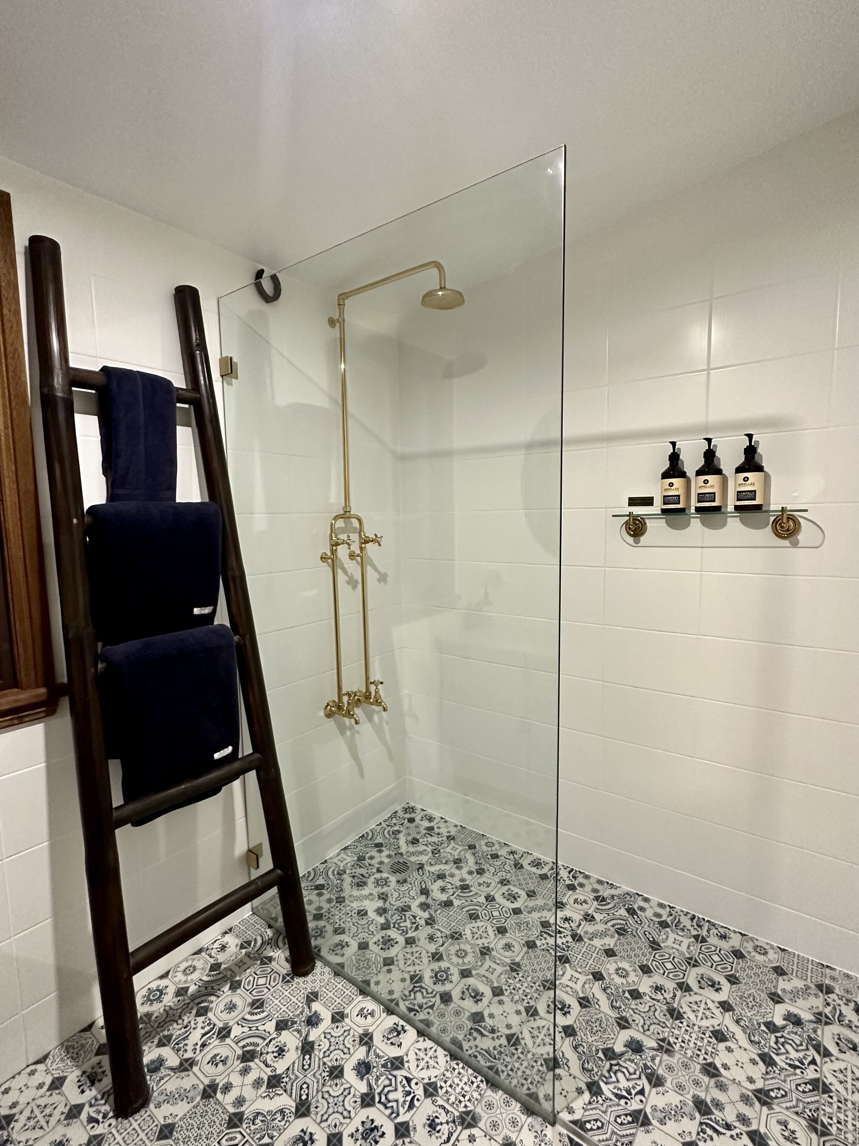 A shower with a glass door, a wooden ladder, and a tiled floor with a geometric pattern.