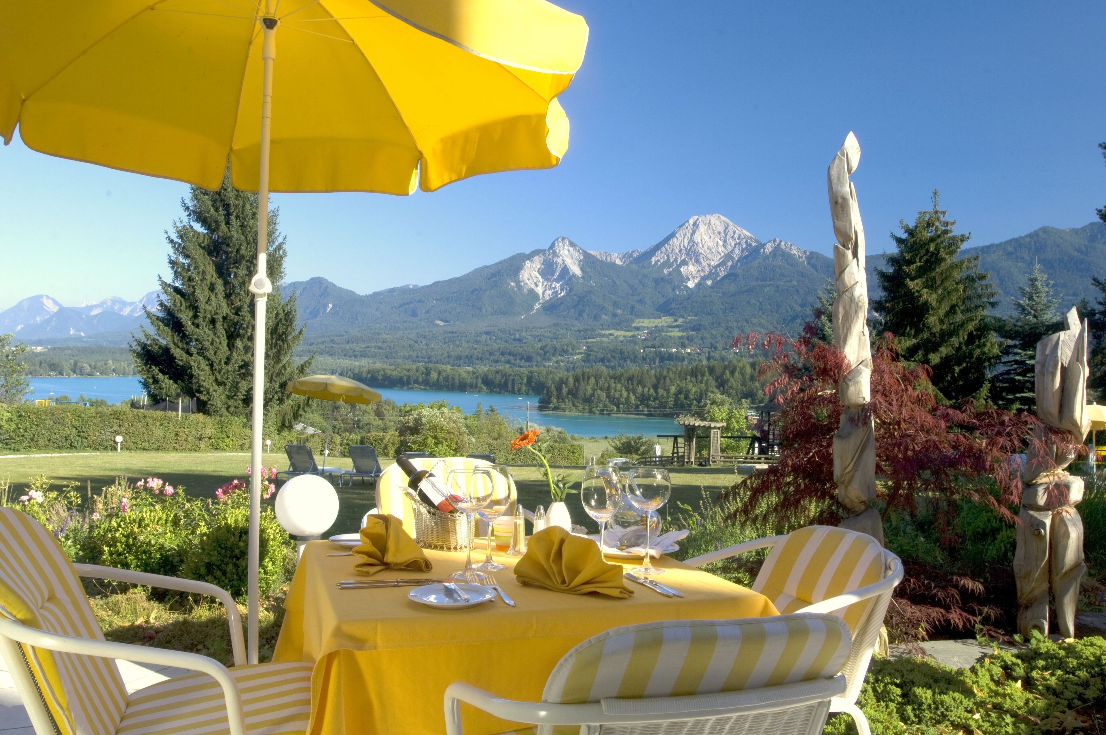 Une table à manger, recouverte d’une nappe jaune et entourée de chaises blanches, offre une vue sur un paysage montagneux avec un lac.