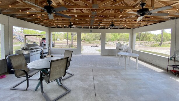 An open-plan indoor space with a concrete floor, wooden ceiling beams, ceiling fans, and large windows offering views of the outdoors.