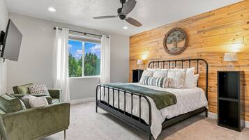 Een slaapkamer met een bed, een groene fauteuil, een houten wand, een plafondventilator en een raam met uitzicht op bomen.