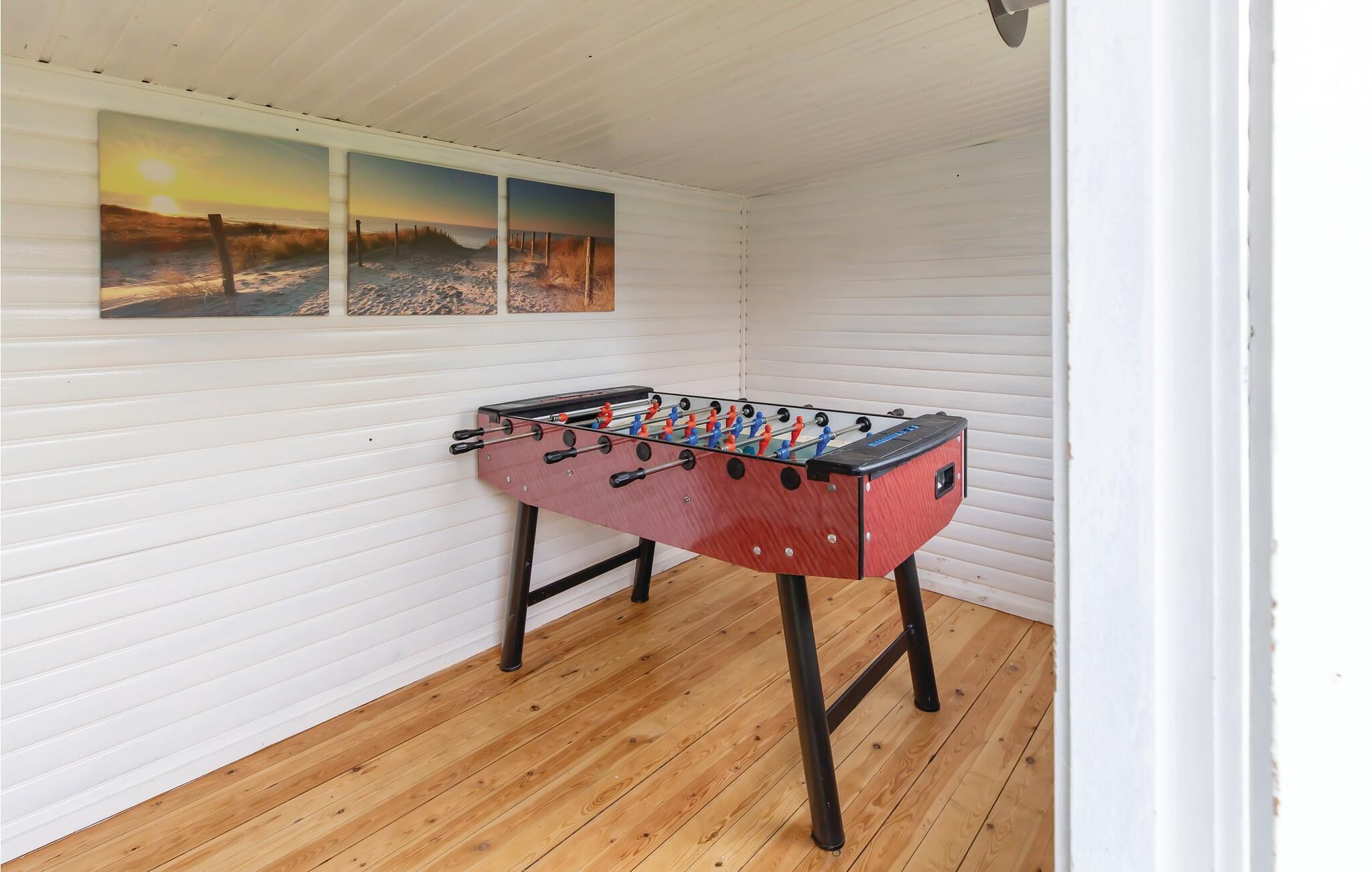 Een kamer met een tafelvoetbaltafel, houten vloeren en drie strandzonsondergangschilderijen aan de muur.