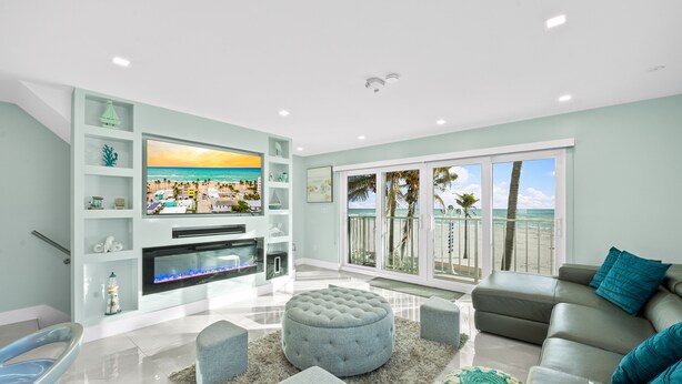 Ein modernes Wohnzimmer mit großem Flachbildfernseher, bequemen Sitzgelegenheiten und Blick auf den Strand durch große Fenster.