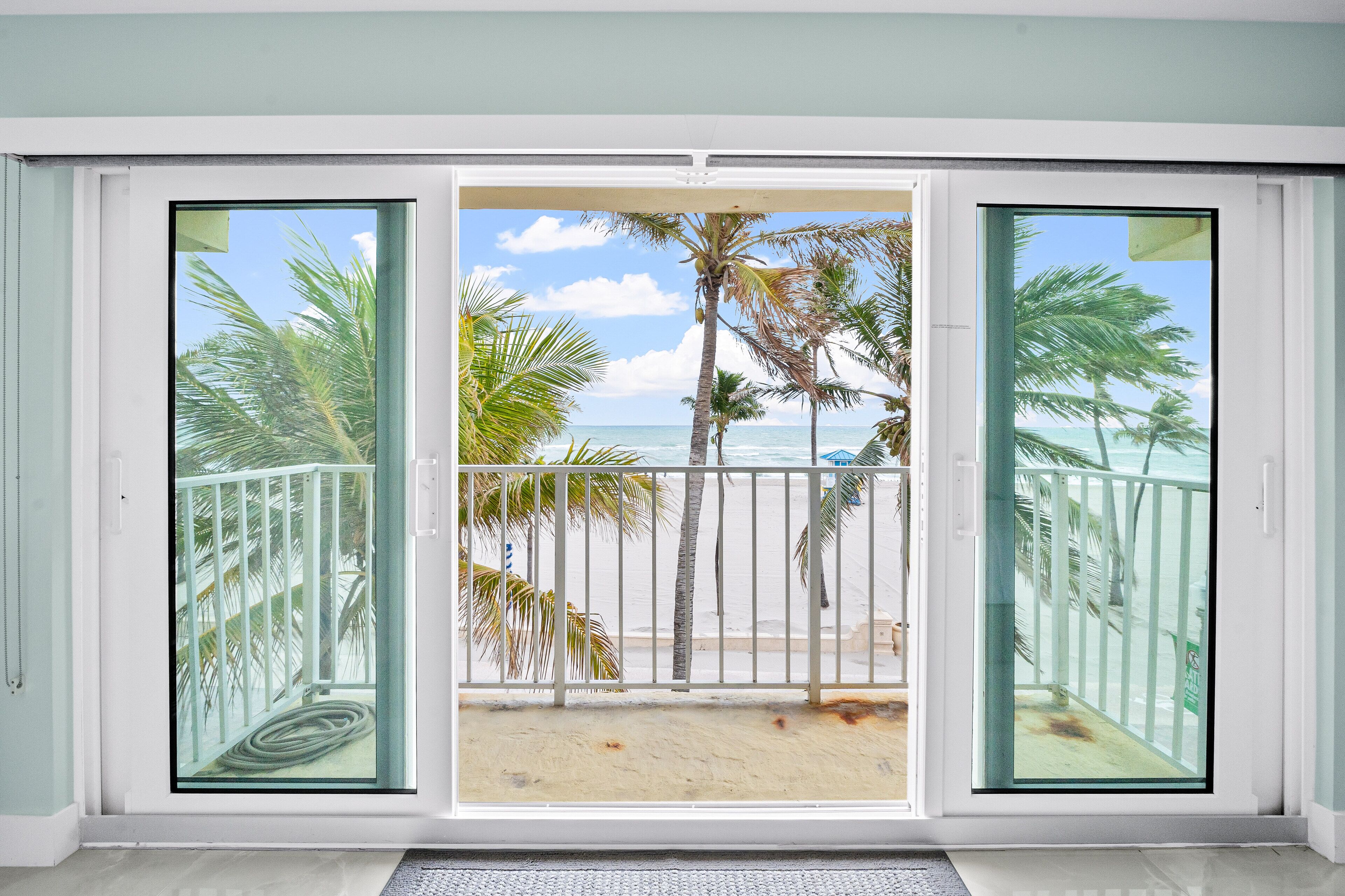 Quarto com uma grande porta de vidro deslizante que dá para uma varanda com vista para uma praia e palmeiras.