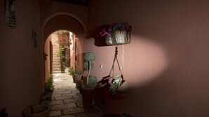 A narrow hallway with a stone floor, leading to a doorway. There are potted plants and a hanging basket with flowers on the right side. - the flowery courtyard srl no12 (Trapani)