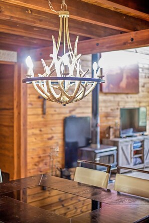 Uma sala de jantar com mesa e cadeiras de madeira, um candelabro com design exclusivo e uma televisão ao fundo.