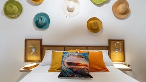 Een bed met een wit kussen en een oranje kussen, twee nachtkastjes en een muur met zeven hoeden.