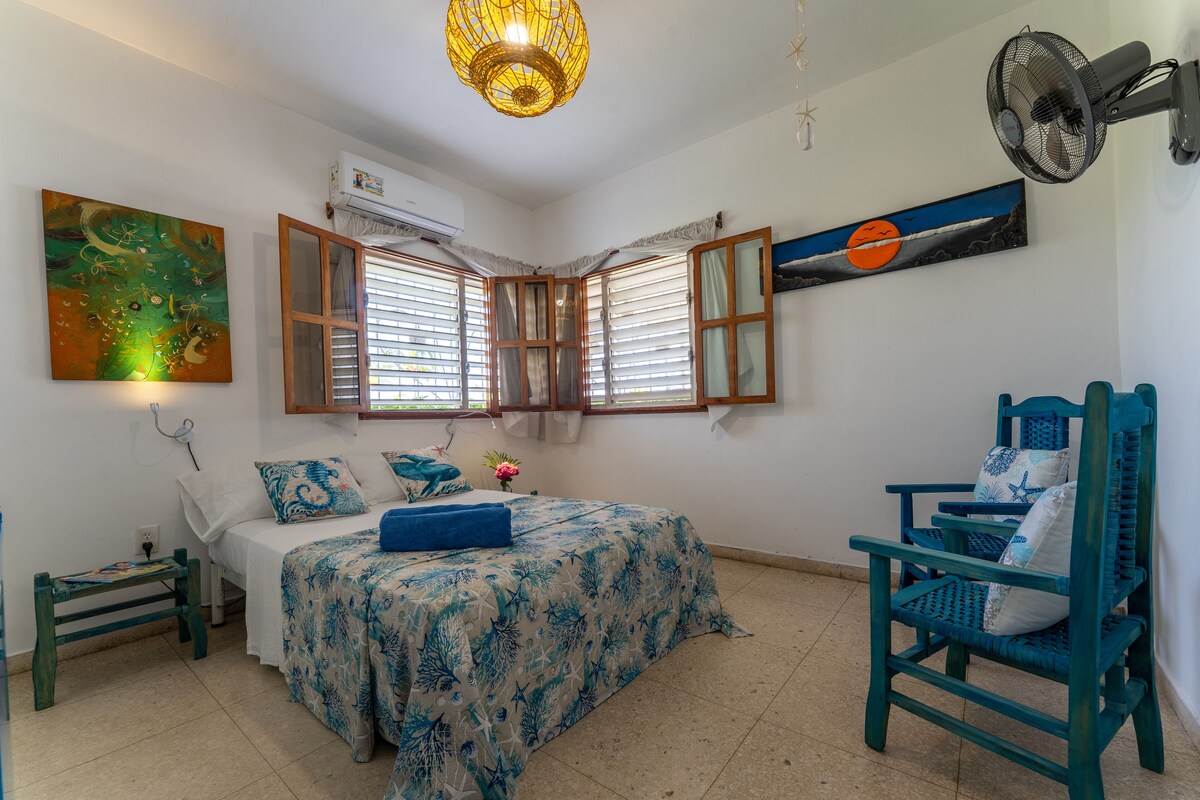 A bedroom with a bed, a chair, a fan, and artwork on the walls.